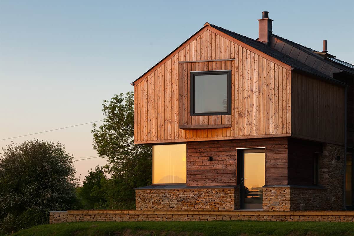 The Larch House timber-clad Passivhaus dwelling with stone base lit warmly at dusk
